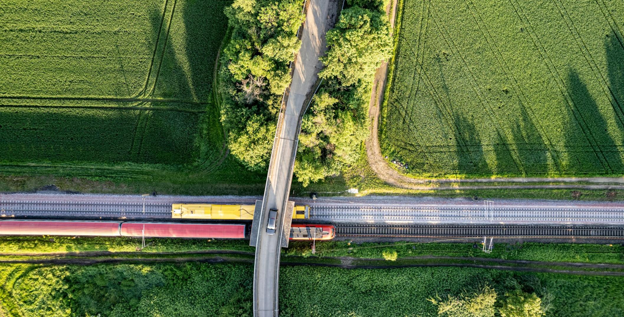  It shows a train crossing a bridge over railway tracks that cut through vibrant green fields. The neat crop rows are contrasted by the curving road and linear railway, illustrating the coexistence of nature and engineered environments. This perspective emphasizes the integration of rural and industrial elements.