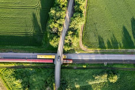  It shows a train crossing a bridge over railway tracks that cut through vibrant green fields. The neat crop rows are contrasted by the curving road and linear railway, illustrating the coexistence of nature and engineered environments. This perspective emphasizes the integration of rural and industrial elements.